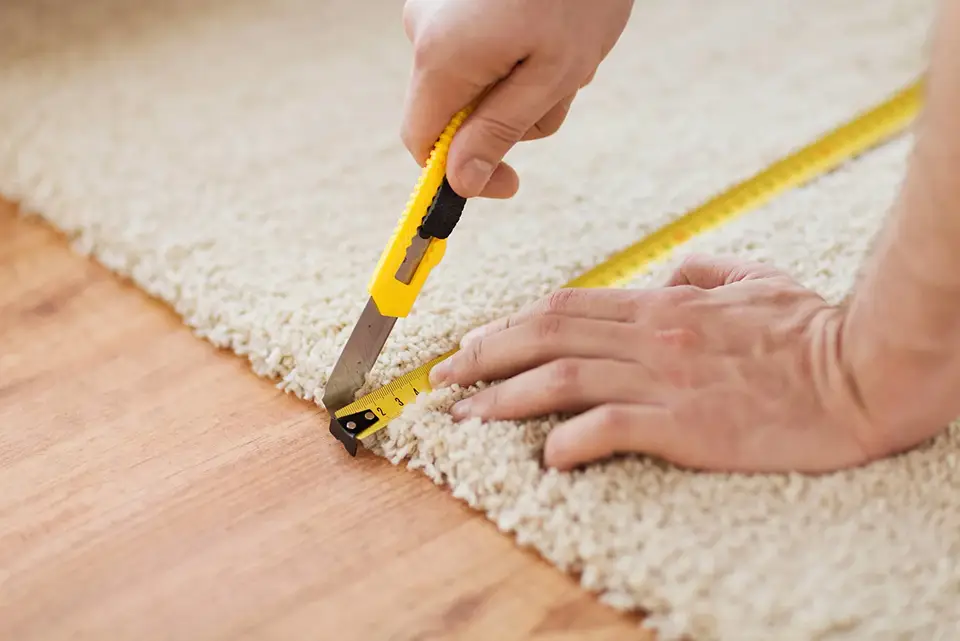 A worker using a utility knife and measuring tape to precisely cut a plush carpet.